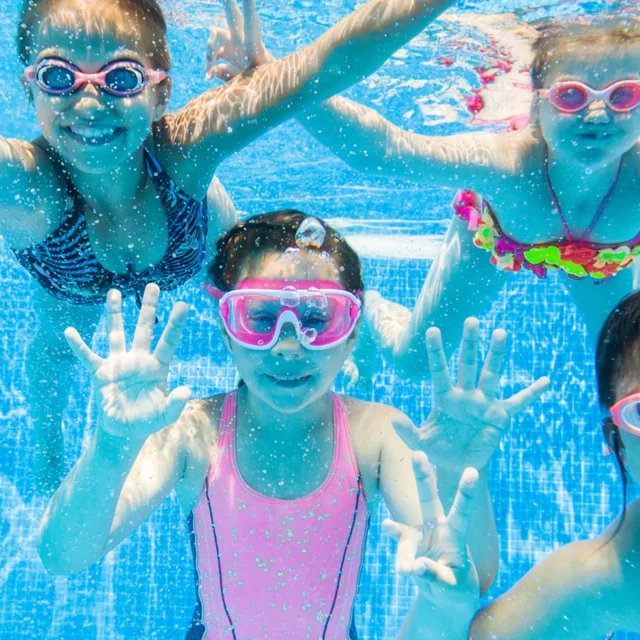 Fünf Kinder, die unter Wasser mit bunten Schwimmbrillen in die Kamera lächeln.