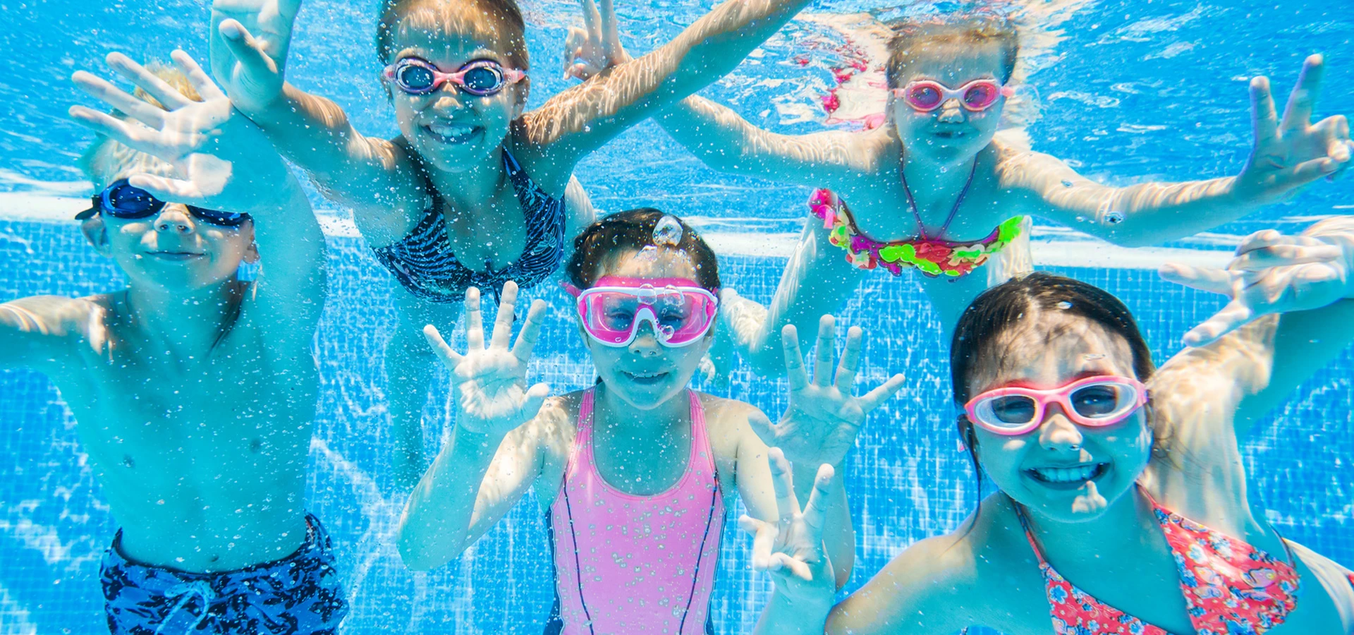 Fünf Kinder, die unter Wasser mit bunten Schwimmbrillen in die Kamera lächeln.