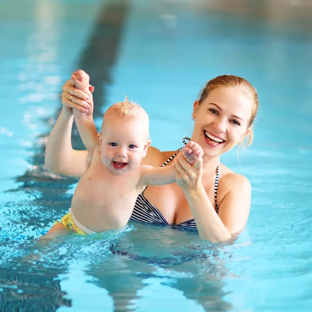 Eine Mutter, die ihr lächelndes Baby an den Händen bis zum Bauchnabel aus dem Wasser hebt.