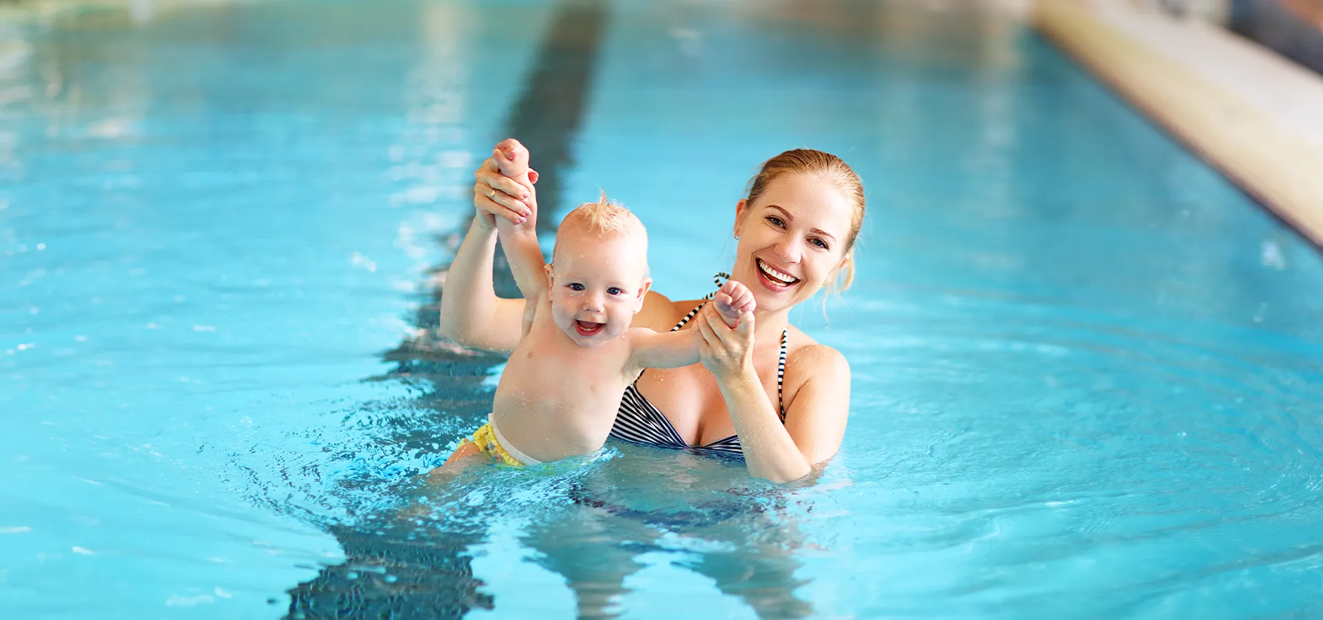 Eine Mutter, die ihr lächelndes Baby an den Händen bis zum Bauchnabel aus dem Wasser hebt.