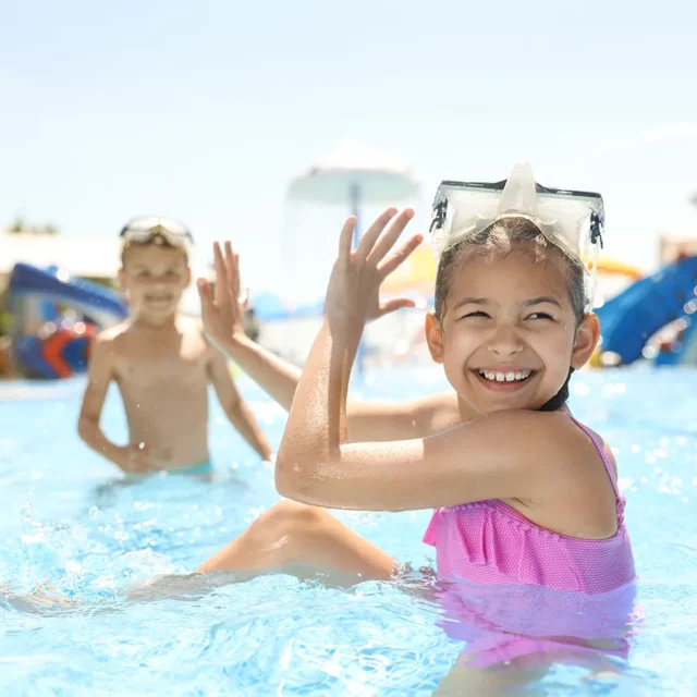 Beim Spielen im niedrigen Wasser versteckt sich das im Vordergrund sitzende Mädchen hinter ihren Händen, um nicht vom Jungen, der im Hintergrund zu erkennen ist, mit Wasser bespritzt zu werden.