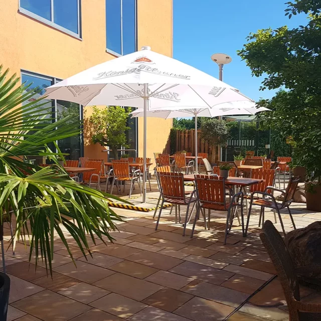 Die Terrasse des Saunarestaurants mit gemütlichen Holztischen und Stühlen im Saunagarten mit ausgebreiteten Sonnenschirmen bei strahlendem Sonnenschein