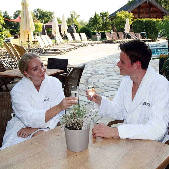 Ein Pärchen hat auf unserer Terrasse am Saunarestaurant Platz genommen und stößt mit einem Glas Sekt an.