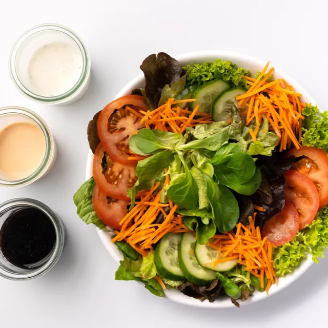 Ein gemischter Salat mit Gurken, Tomaten, Karottenstreifen und drei verschiedenen Dressings in separaten Glasschälchen von oben auf weißem Untergrund.