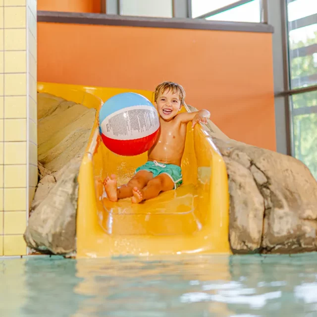 Ein lachender Junge beim Rutschen im Kinderbecken mit einem Wasserball in der Hand