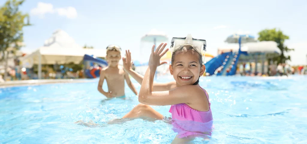 Beim Spielen im niedrigen Wasser versteckt sich das im Vordergrund sitzende Mädchen hinter ihren Händen, um nicht vom Jungen, der im Hintergrund zu erkennen ist, mit Wasser bespritzt zu werden.