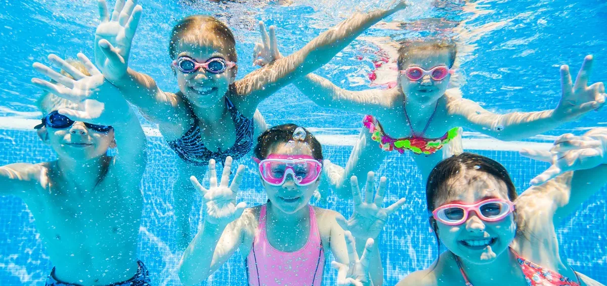 Fünf Kinder, die unter Wasser mit bunten Schwimmbrillen in die Kamera lächeln.