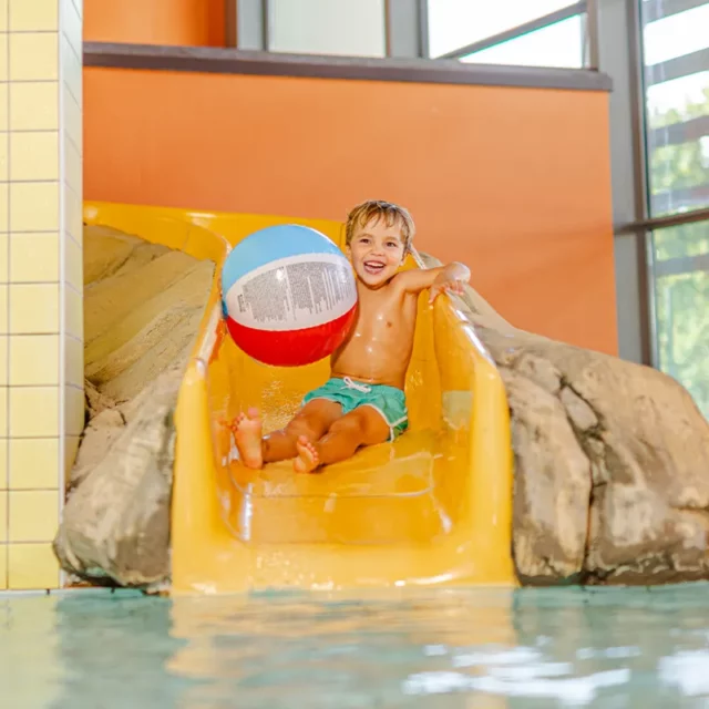 Ein lachender Junge beim Rutschen im Kinderbecken mit einem Wasserball in der Hand