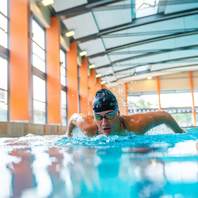 Die Schwimmerin taucht beim Schmetterlingsschwimmen aus dem Wasser auf. Sie trägt Badekappe und Schwimmbrille.